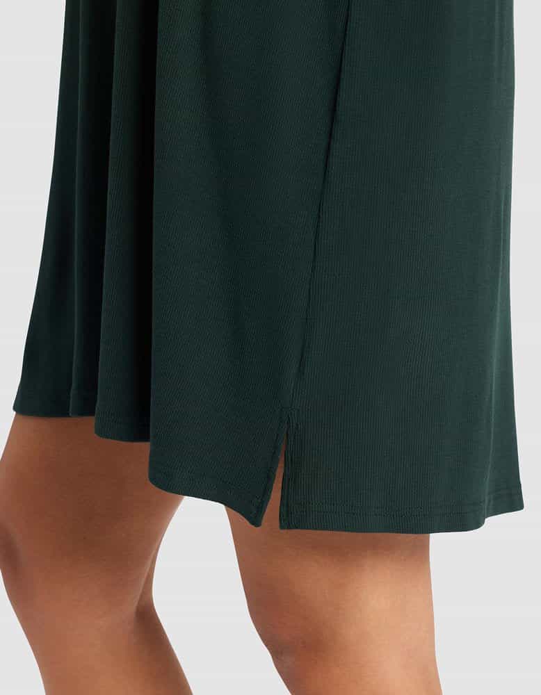 A close-up of a person wearing the Cozy Earth Women’s Bamboo Rib-Knit Boyfriend Sleep Dress in dark green, featuring a small side slit that reveals part of their upper leg against a plain, light background.|Color:Spruce