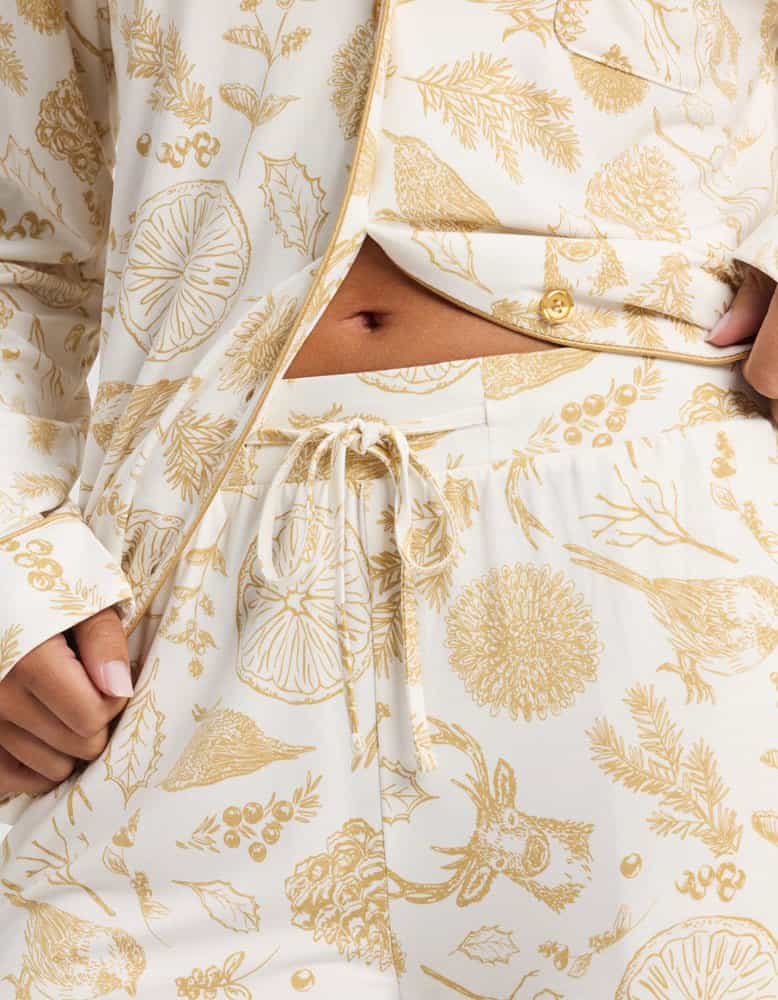 A person wears Cozy Earth’s Women's Bamboo Stretch-Knit Pajama Pant in cream, featuring golden botanical and animal prints, a visible drawstring, and a partially unbuttoned top that reveals the stomach.|Color:Winter Toile Dijon