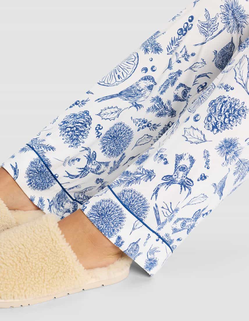 Close-up of a person wearing cream-colored slippers and Cozy Earth Women's Bamboo Stretch-Knit Pajama Pant, featuring a blue nature print with birds, leaves, pinecones, and acorns, set against a light background.|Color:Winter Toile Pacific
