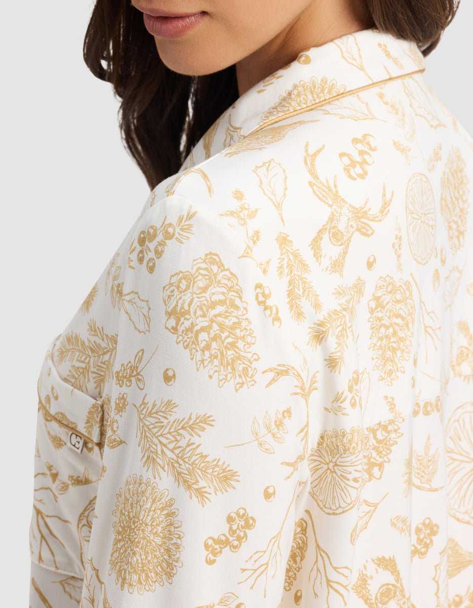 A woman wears the Cozy Earth Women's Bamboo Stretch-Knit Long Sleeve Pajama Top, featuring a white base with gold botanical and woodland animal prints—pinecones, sprigs, berries, and a stag's head. Side view highlights the shirt's detailed pattern.|Color:Winter Toile Dijon